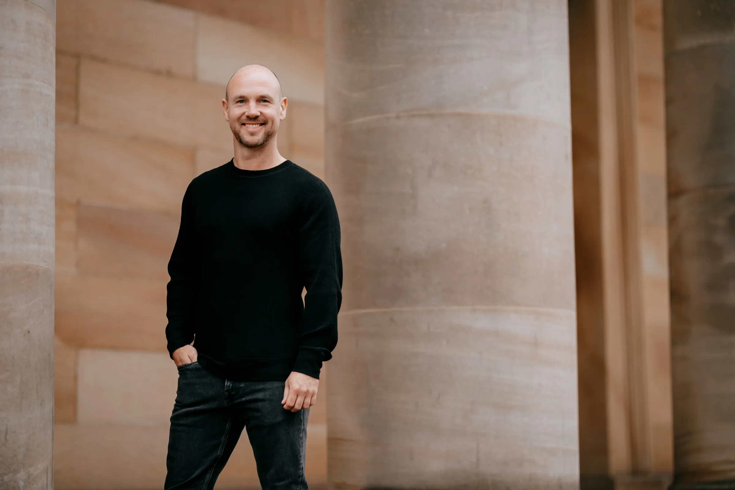 Paul Holmes smiling between sandstone columns, wearing a black jumper.