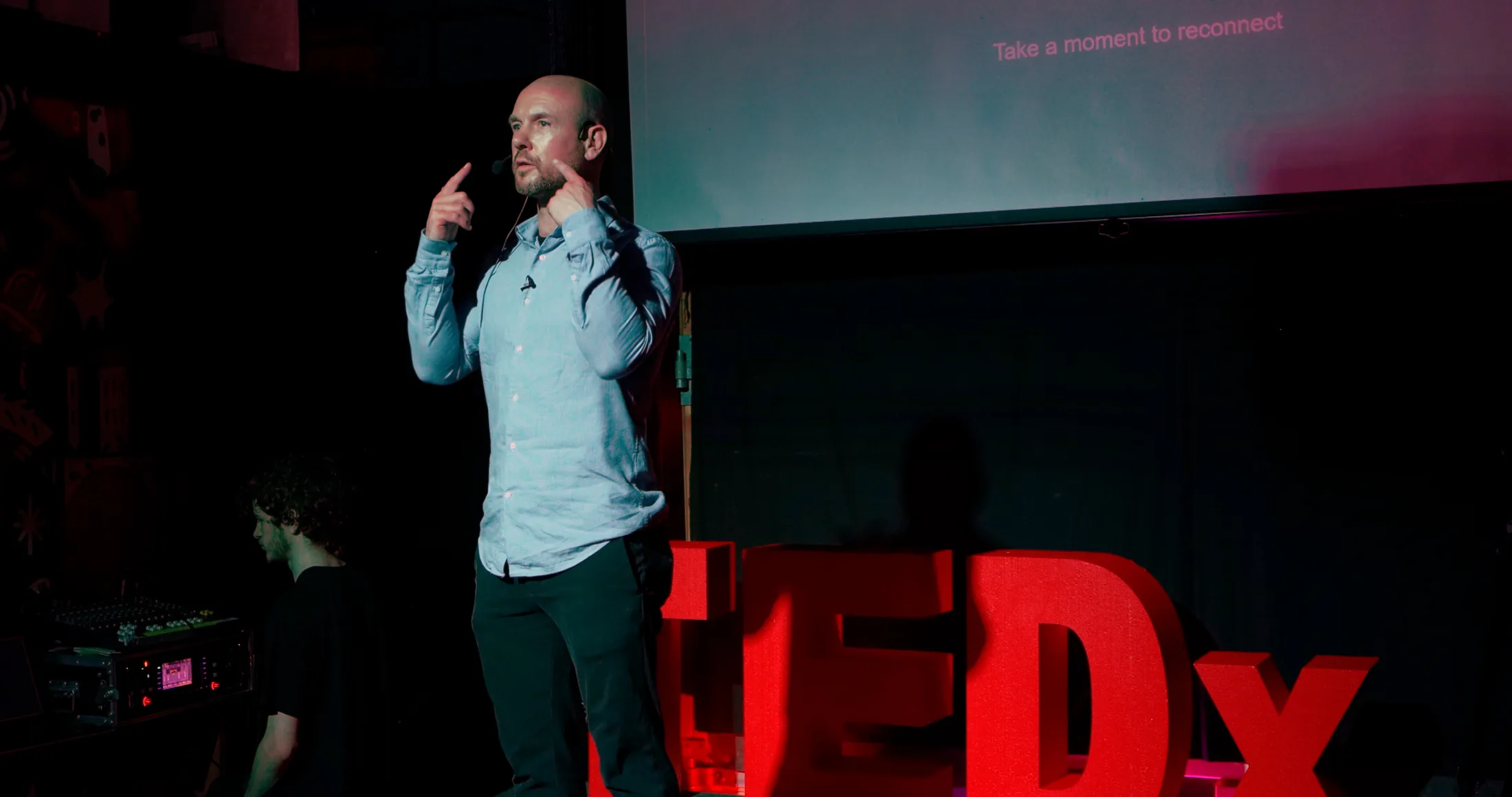 Paul Holmes speaking on a TEDx stage beside the red TEDx letters.